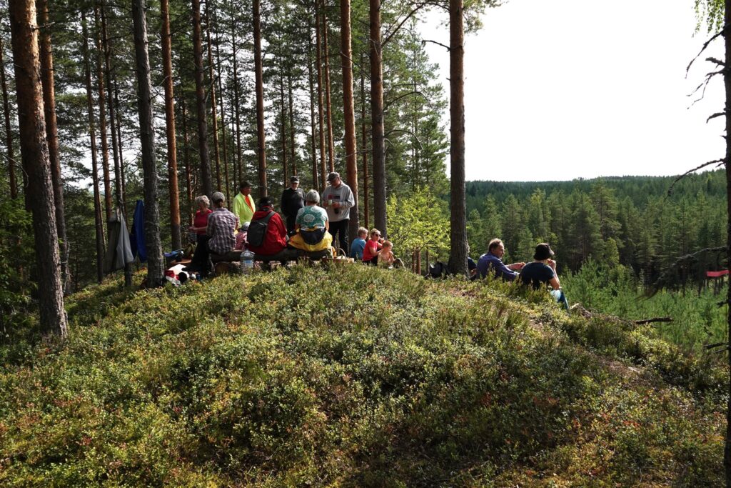 Syrjäharjun luontopolkujen näköalapaikalla kukkulalla istuu retkeilijöitä maassa ja penkeillä syömässä eväitä.