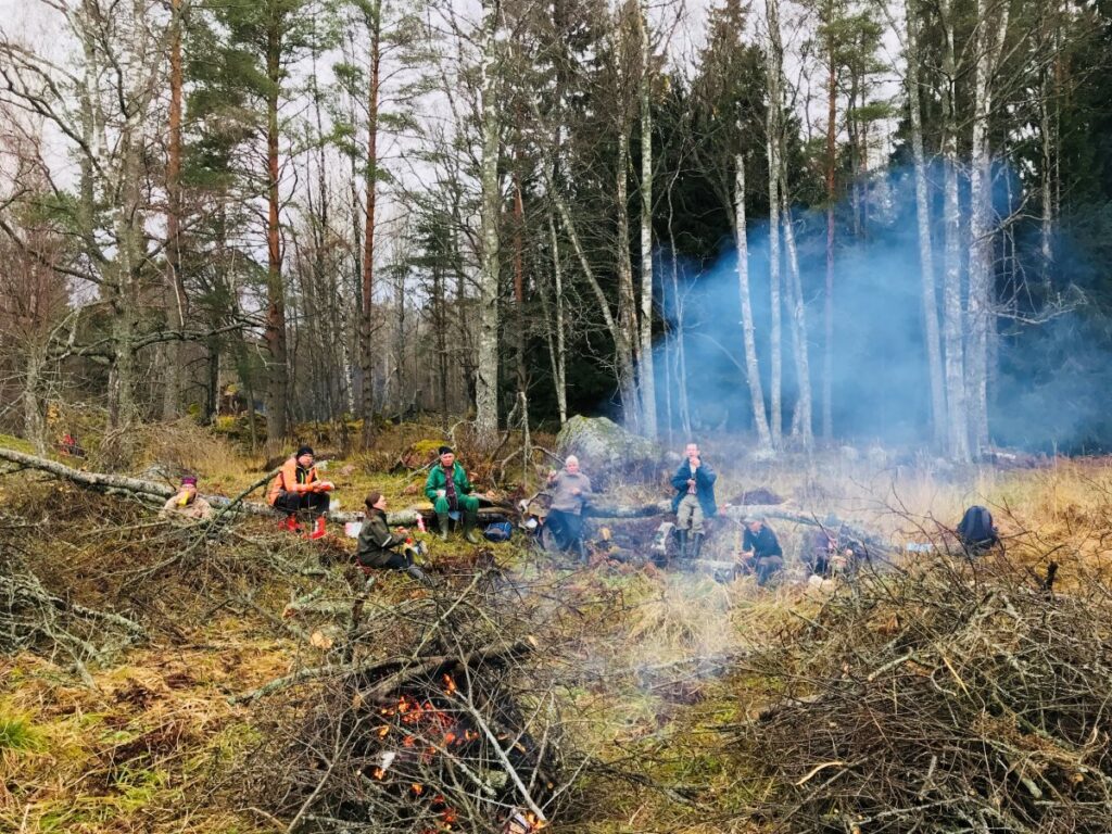 Seitsemän ihmistä istuu marraskuisessa maisemassa evästauolla. Etualalla savuaa poltettava risukasa, taustalla on sekametsää.