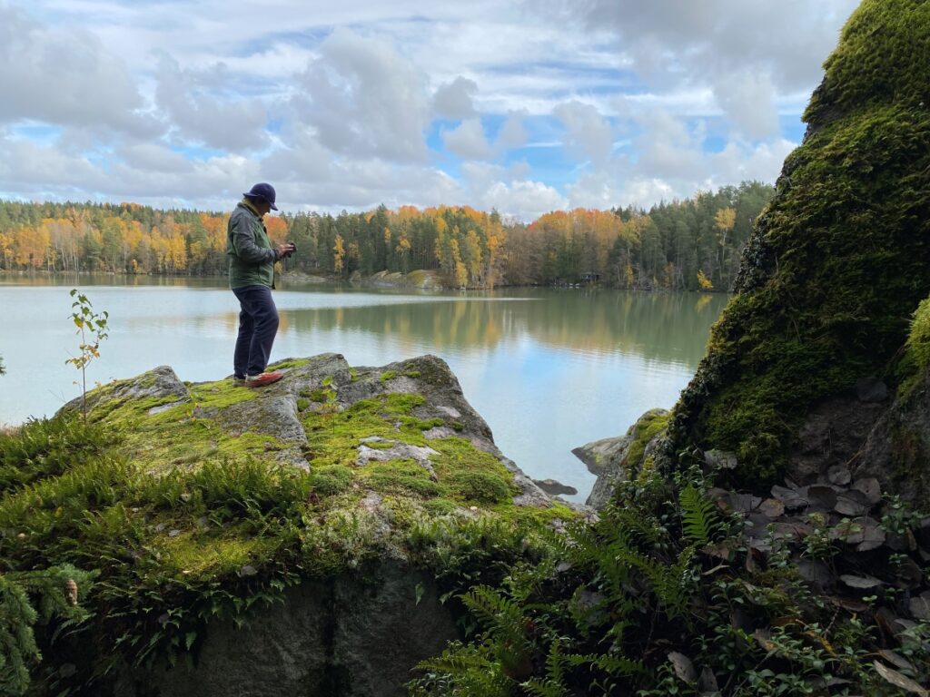 Ihminen kalliolla, taustalla järvi ja metsää. Puolipilvinen taivas.