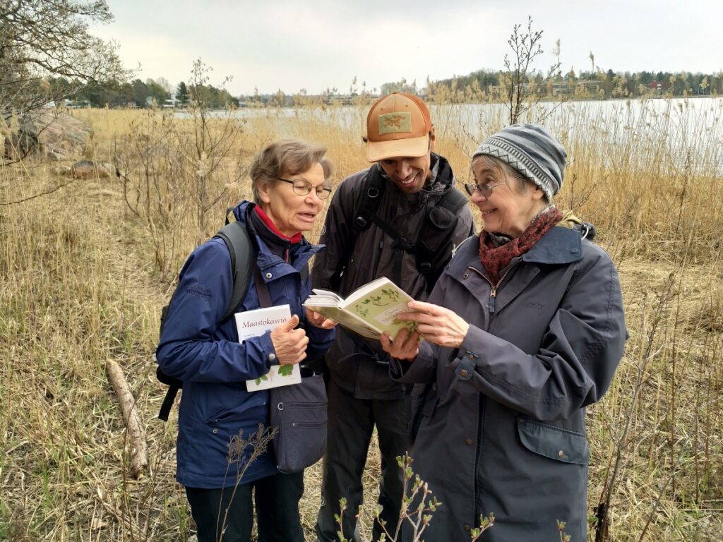 Kolme ihmistä tutkii kasvikirjaa, taustalla ruovikkoa ja merta. 