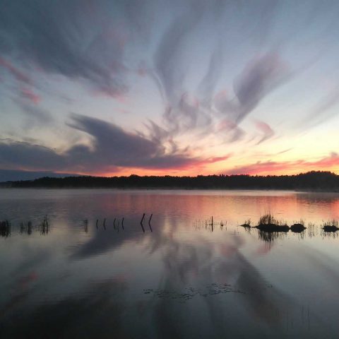 Kuvassa näkyy rauhallinen järvimaisema illalla. Aurinko on laskenut horisontin taakse ja taivaalla on hieman pilviä.