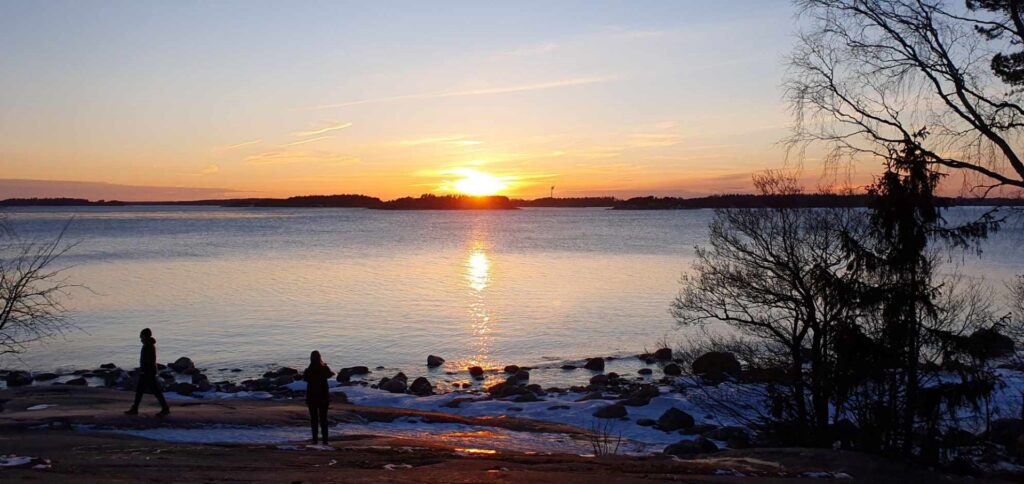 Talvinen auringonlasku meren rannassa, etualalla kalliota ja kaksi tummaa ihmishahmoa sekä tumma lehdetön puunrunko.