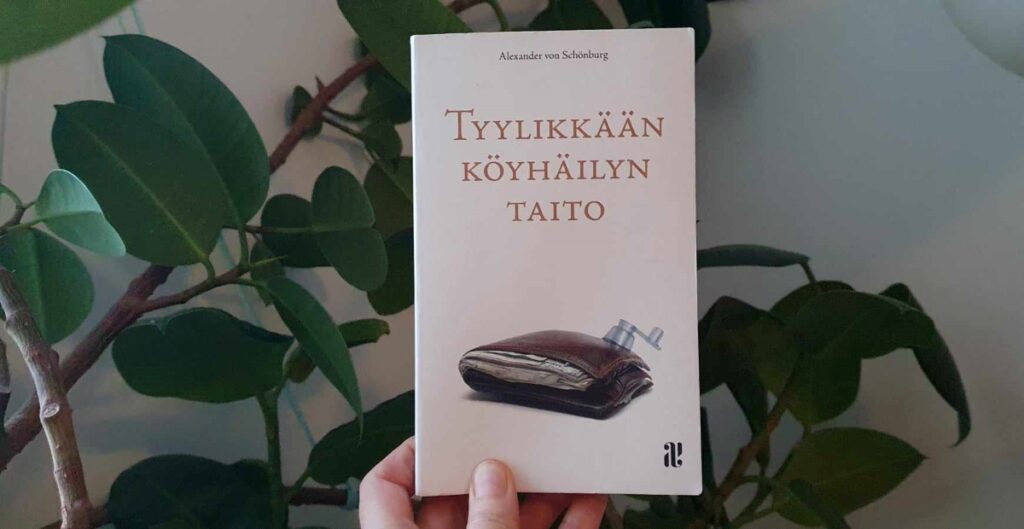 Tyylikkään köyhäilyn taito -kirja ihmisen kädessä, taustalla huonekasvi (kumipuu)