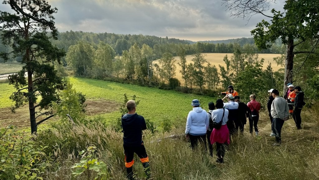 Ihmisiä niityn reunalla katselemassa maisemaa, taustalla metsää, peltoa ja taivasta.