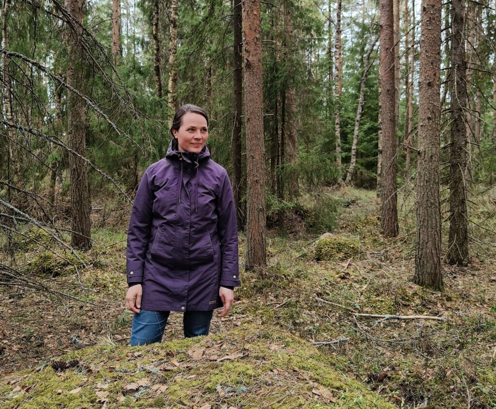 Hanna Halmeenpää seisoo syksyisessä metsässä kuusten ja mäntyjen ympäröimänä ja katsoo sivulle sekä hymyilee.